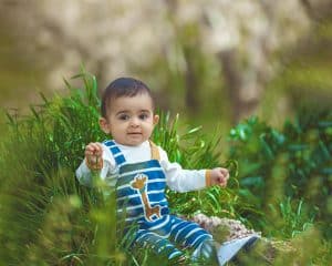 متن جایگزین پیشنهادی: پرتره‌ای از یک نوزاد با چشمانی درشت و کنجکاو که در میان چمن‌های سبز و باطراوت نشسته و مستقیم به دوربین نگاه می‌کند. نوزاد یک لباس سرهمی راه‌راه آبی و سفید با طرح زرافه پوشیده است. پس‌زمینه‌ی محو و طبیعی، تمام توجه را به معصومیت و زیبایی کودک جلب می‌کند.