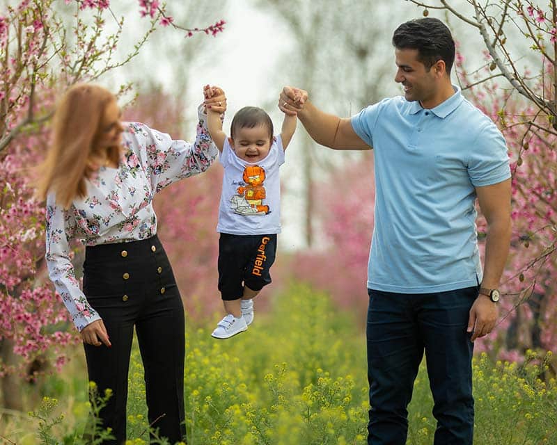 پدر و مادری در یک دشت پر از گل‌های زرد و شکوفه‌های صورتی، دستان کودک خندان خود را گرفته و او را به آرامی در هوا تاب می‌دهند. این تصویر، یک لحظه‌ی بازیگوشانه، شاد و پر از انرژی مثبت خانوادگی در طبیعت زیبا را ثبت کرده است.