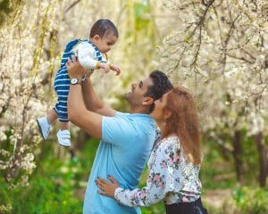 یک پدر و مادر در باغی پر از شکوفه‌های سفید بهاری، با عشق به نوزادشان نگاه می‌کنند. پدر، نوزاد را که لباس راه‌راه آبی و سفید به تن دارد، با دستانش به سوی آسمان بلند کرده و مادر با لبخند این صحنه را تماشا می‌کند. این عکس، لحظه‌ای سرشار از محبت و شادی یک خانواده را به تصویر می‌کشد.