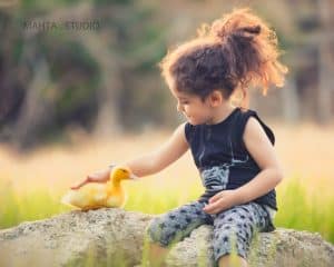 کودک با موهای فرفری و لباس تیره در حال نوازش یک جوجه اردک زرد در فضای باز، عکس برند شده توسط آتلیه مهتا.