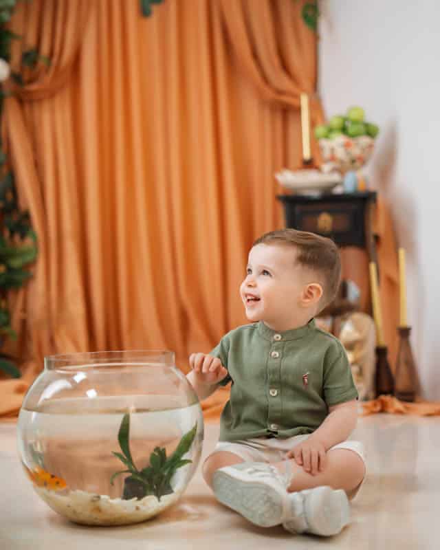 پسر بچه ای در فضای هفت سین نوروزی آتلیه کودک کنار تنگ ماهی نشسته و میخندد