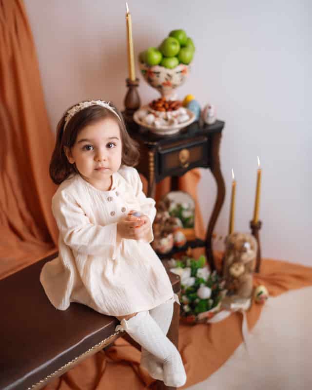 کودک خندان با لباس نخی کرم و جوراب بلند روی نیمکت در دکوراسیون آتلیه برای نوروز